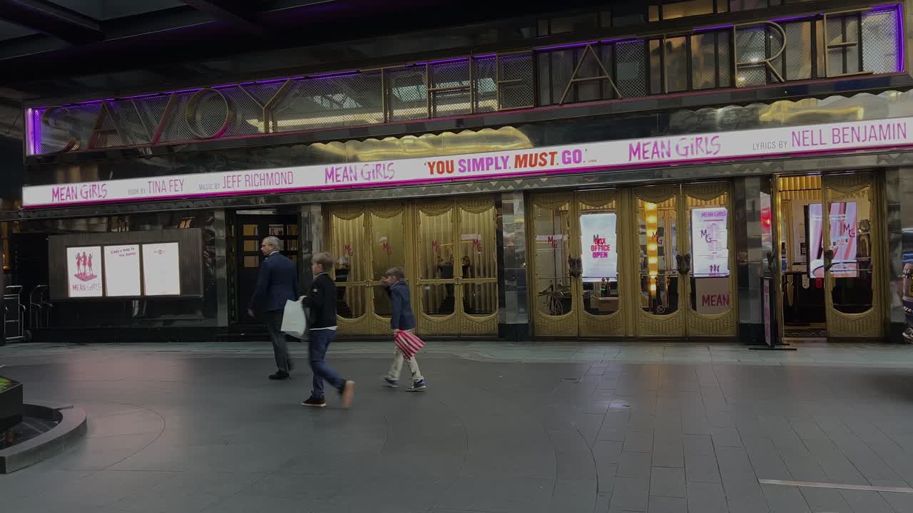 Londoners walk by the entrance of the Savoy Theatre, which features signage for Mean Girls: The Musical. Capturing the energy of London's West End and the theater's vibrant appeal
