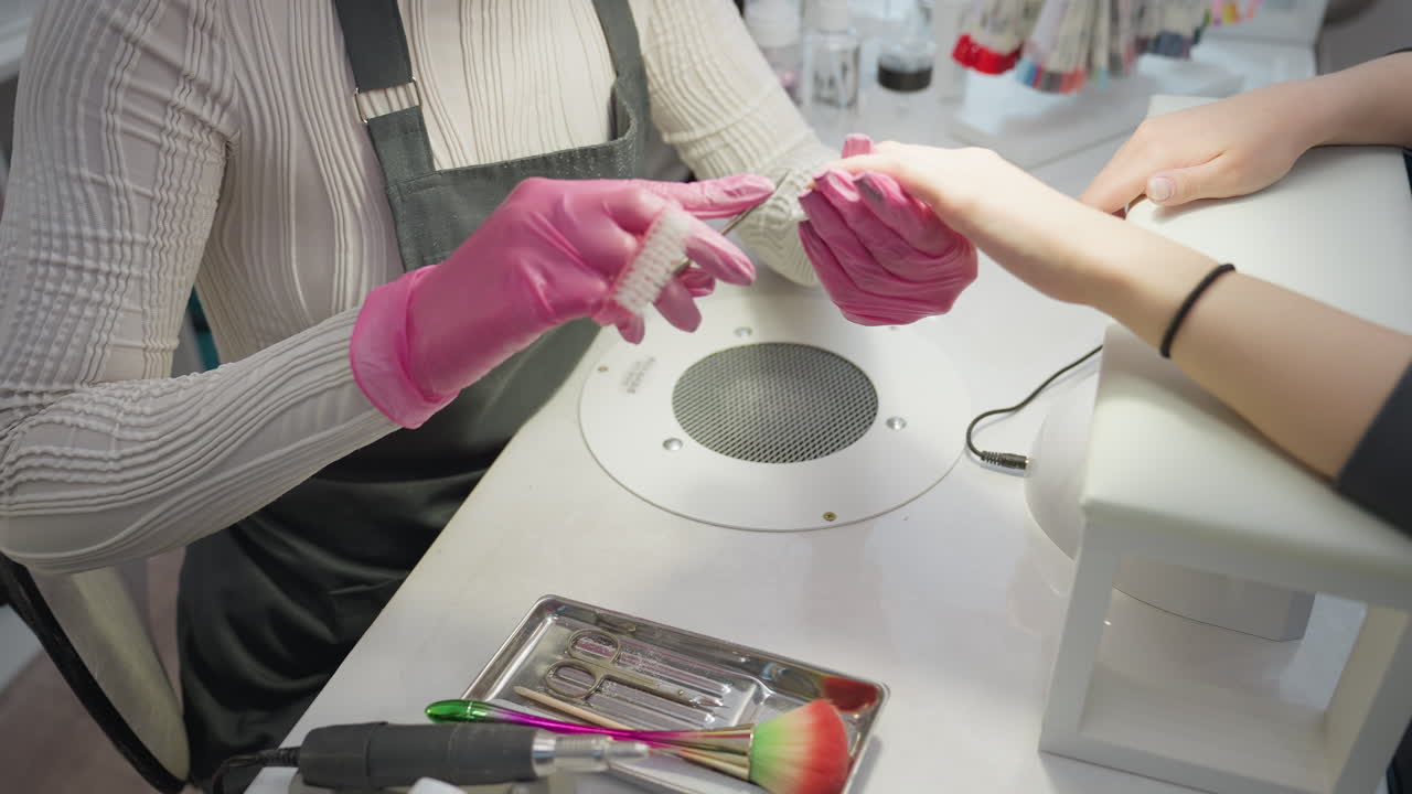 Nail technician in pink gloves carefully using scissors and brush on customer nails while seated at manicure table with tools and bottles, focusing on grooming and hygiene in clean salon setting