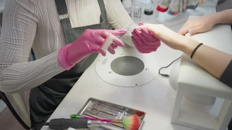 nail technician in pink gloves carefully using scissors and brush on customer nails while seated at manicure table with tools and bottles, focusing on grooming and hygiene in clean salon setting