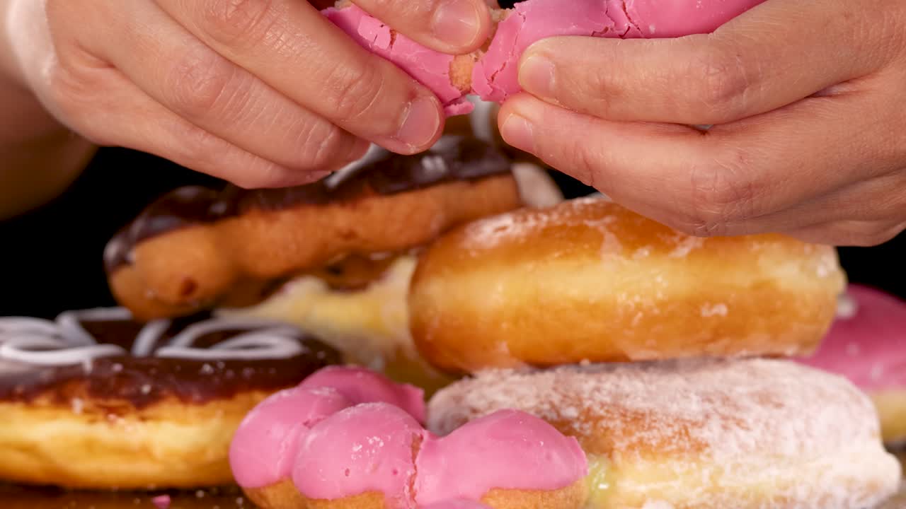 Hands break a pink glazed donut over assorted donuts under bright studio lighting, black background
