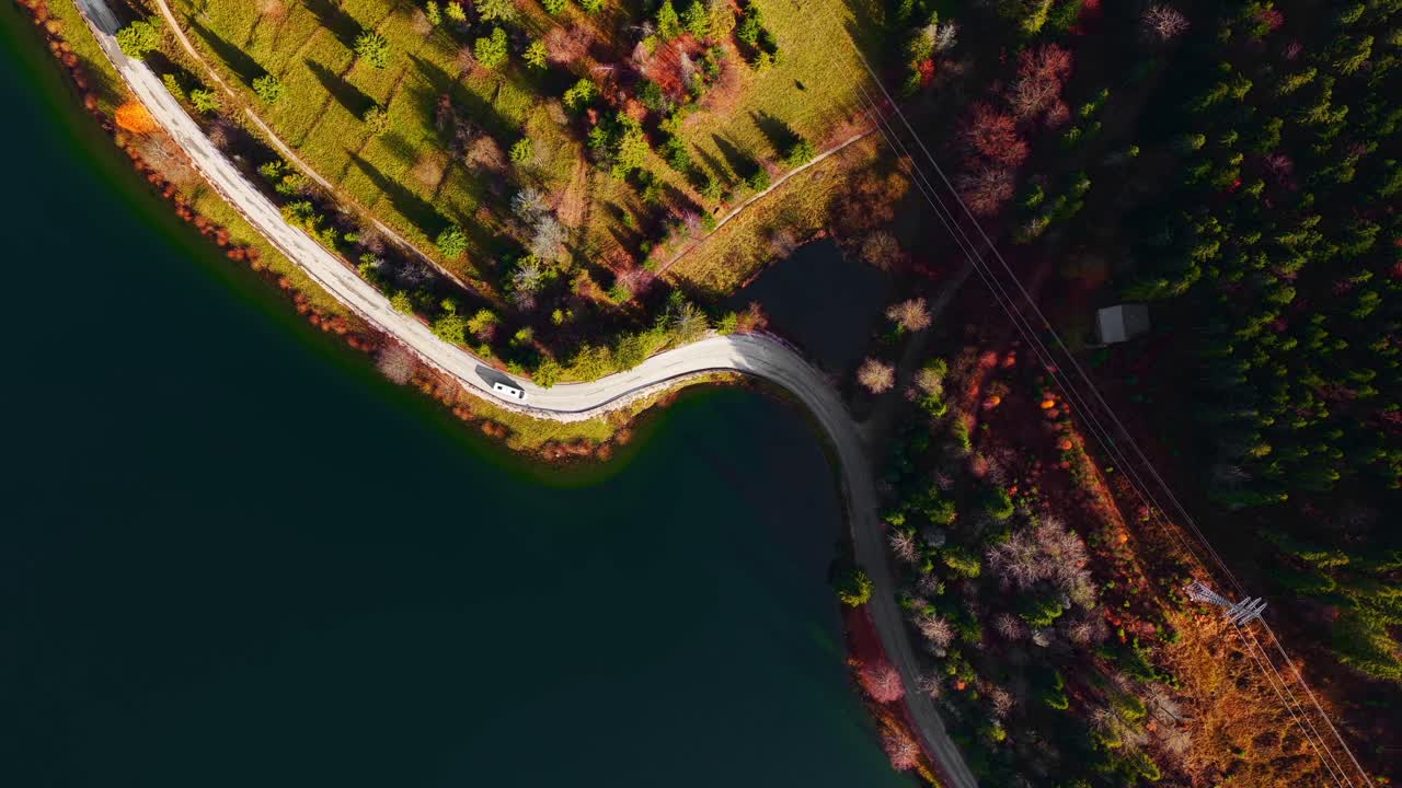 Drone view of a winding road along a calm lakeshore, bordered by lush green hills, trees, and warm autumn tones in a peaceful natural setting