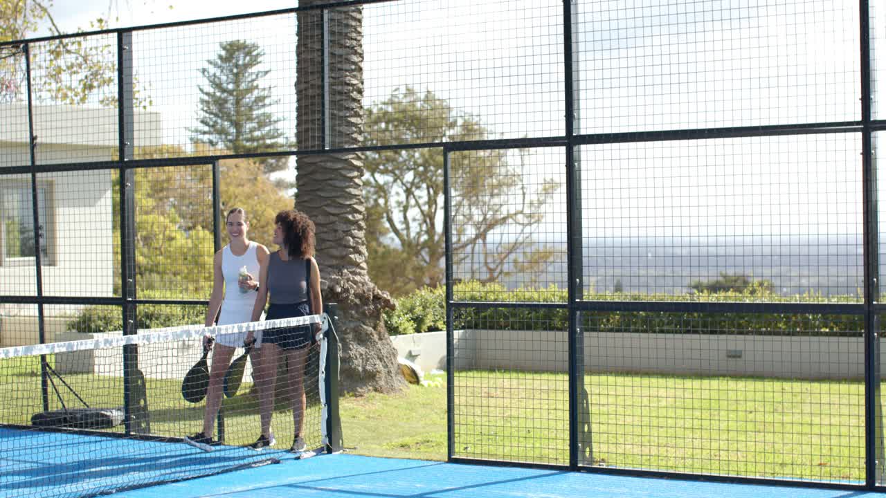 Diverse amiche camminano su un campo da padel blu portando racchette e borse per giocare.