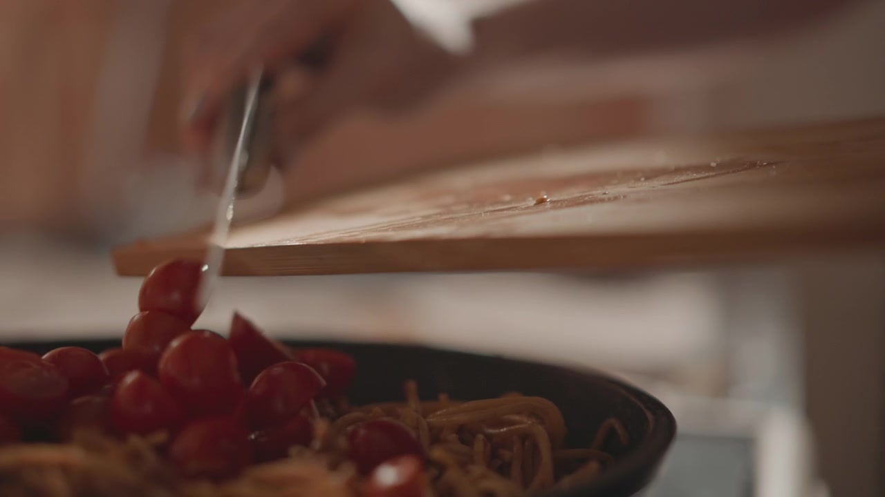 Vue rapprochée de tomates cerises coupées en tranches glissant d'une planche à découper en bois et tombant dans une poêle chaude de nouilles et de légumes cuits, avec des éclaboussures de jus, de la vapeur s'élevant et un mouvement visible