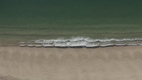 Eine-Aufnahme-Von-Oben-Nach-Unten,-In-Der-Kleine-Wellen-Einen-Goldenen-Sandstrand-Umspülen