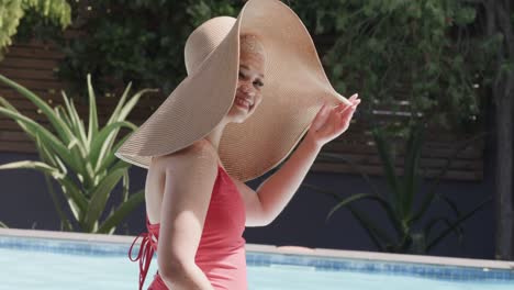 Portrait-of-happy-biracial-woman-with-sun-hat-at-swimming-pool-in-slow-motion