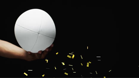 Animation-of-confetti-over-white-rugby-ball-held-by-caucasian-man's-hand-on-black-background