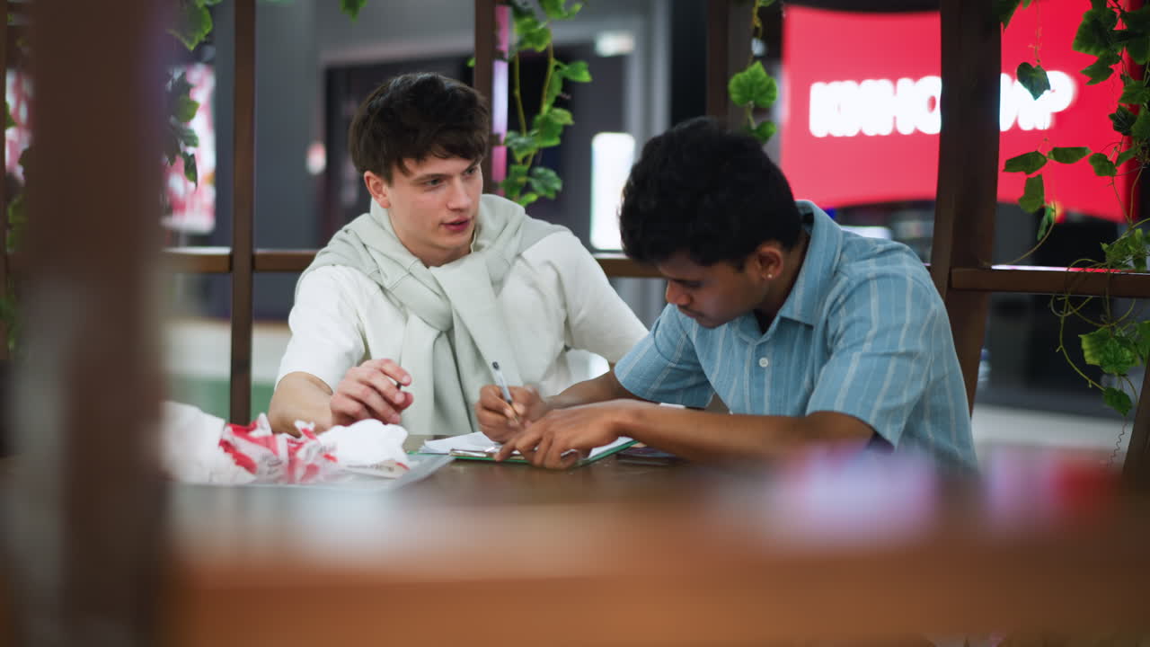 Moving shot capturing two friends drawing sketches by wooden pillars outdoors while vivid television screen displays imagery overhead beams cast on floor evoking creative outdoor collaboration vibe