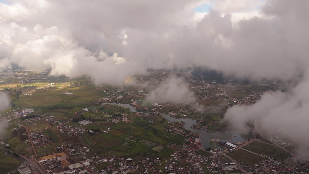 High-altitude drone view over Chignahuapan, flying above scattered low clouds and revealing fields, neighborhoods and distant urban structures beneath