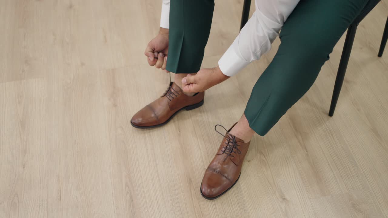 Man in green pants tying classic brown leather shoes on light wooden floor before a wedding