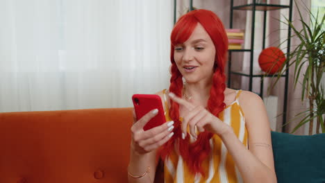 cheerful ginger woman sitting on sofa, using smartphone share messages on social media application