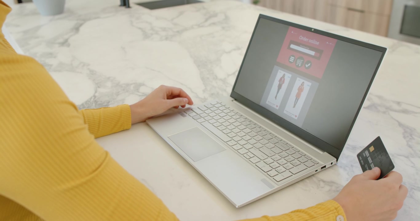 Mulher de blusa amarela na ilha da cozinha finalizando a compra no laptop, digitando, segurando o cartão de crédito para pagar.