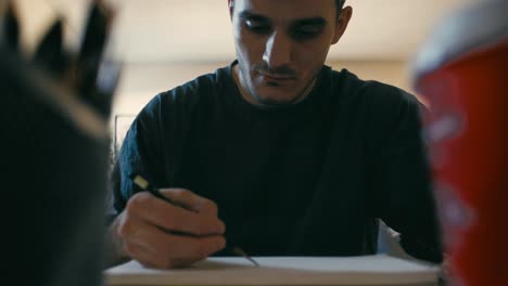 Camera-frames-an-artist-sketching-on-his-pad,-nestled-between-an-out-of-focus-coffee-cup-and-pencil-holder