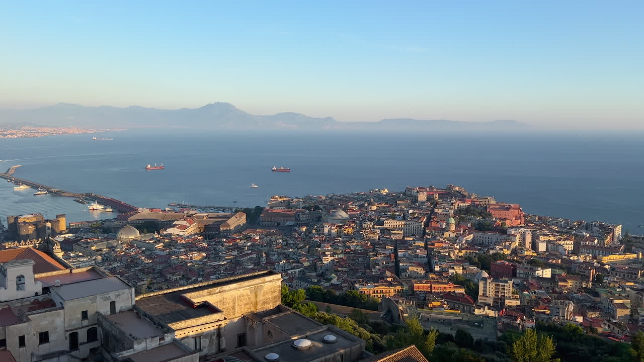 Elevated view of Naples with a focus on the city's dense architecture, the coastline, and the glistening Mediterranean Sea at sunrise