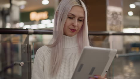 reflective woman seated on bench in bright mall corridor wearing white knit sweater and jeans, focused on tapping tablet screen, black handbag by side, modern glass railing