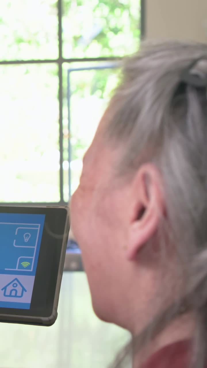Video verticale: Donna anziana asiatica con un top bordeaux che chiude la porta toccando un tablet smart in cucina.