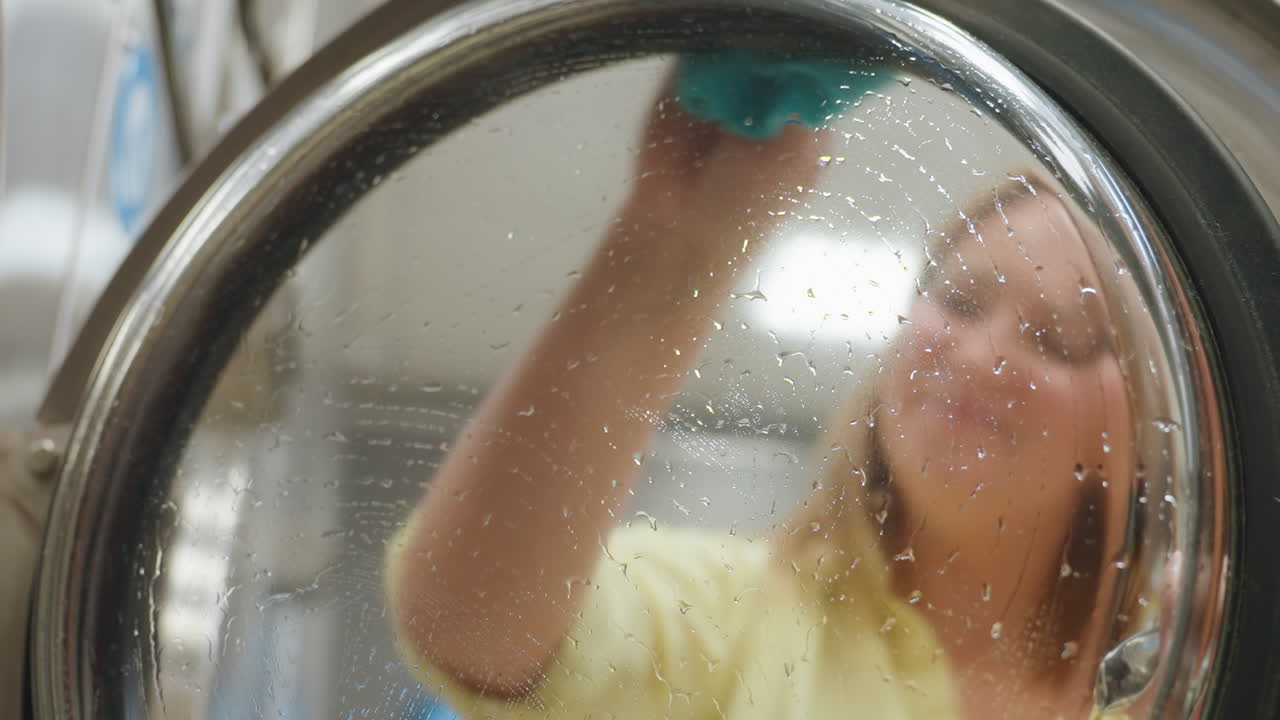 Excited woman seen through transparent washing machine door cleaning surface with wet towel, droplets visible on glass as she enjoys cleaning moment inside laundromat with enthusiasm