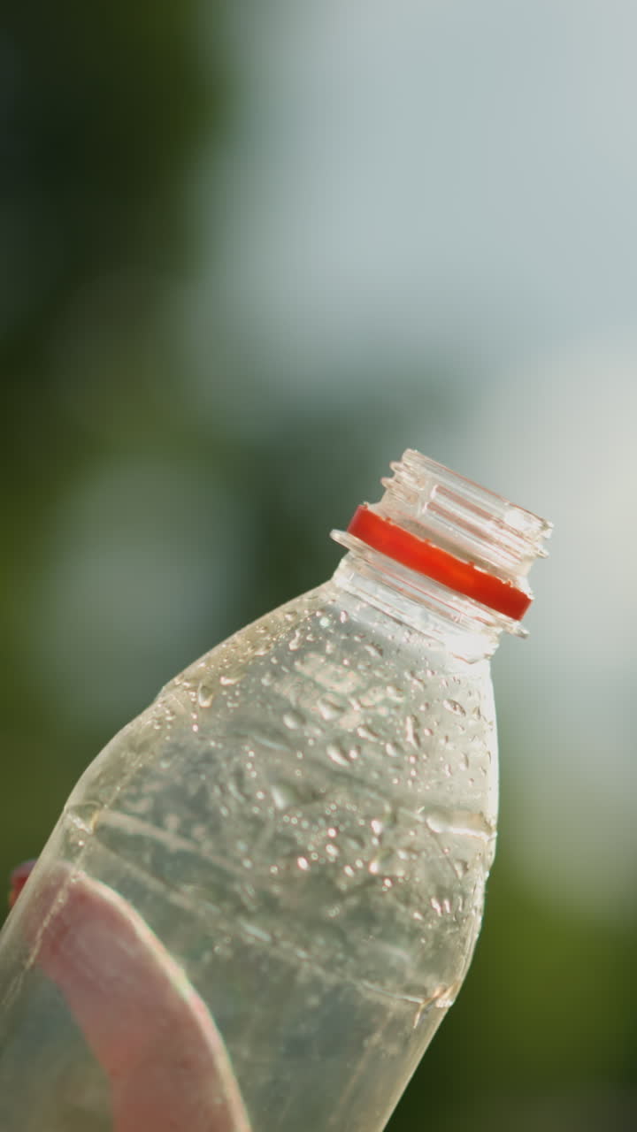 내부에 응축물이 들어있는 투명한 플라스틱 병을 들고 있는 손의 클로즈업, 병은 빛을 향해 기울어져 있으며, 빛은 배경에 부드러운 빛을 일으키며, 흐릿한 초록색으로 둘러싸여 있습니다.