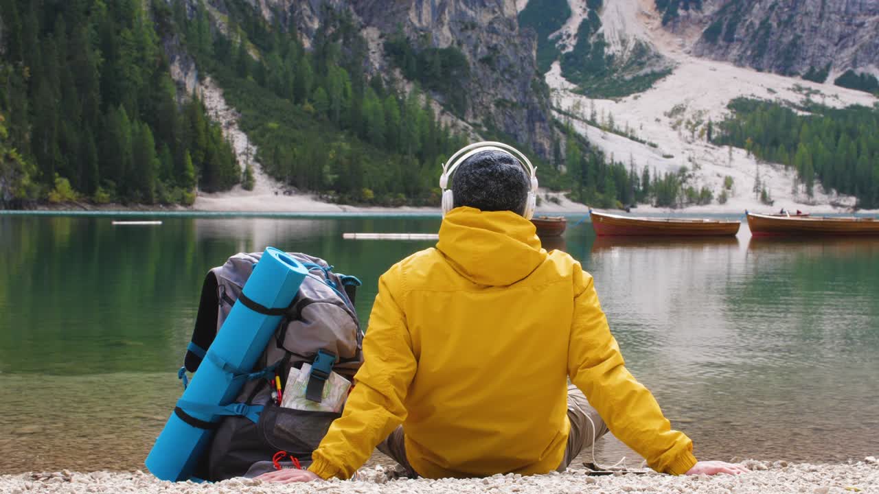 Giovane escursionista turista che ascolta musica seduto vicino al lago alpino Lago di Braies (Pragser Wildsee) in Trentino, Dolomiti, Italia