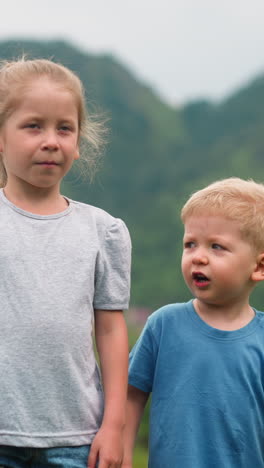 Portrait of cute little siblings boy and girl in casual clothes standing on field against large old forestry hills on cloudy day close view