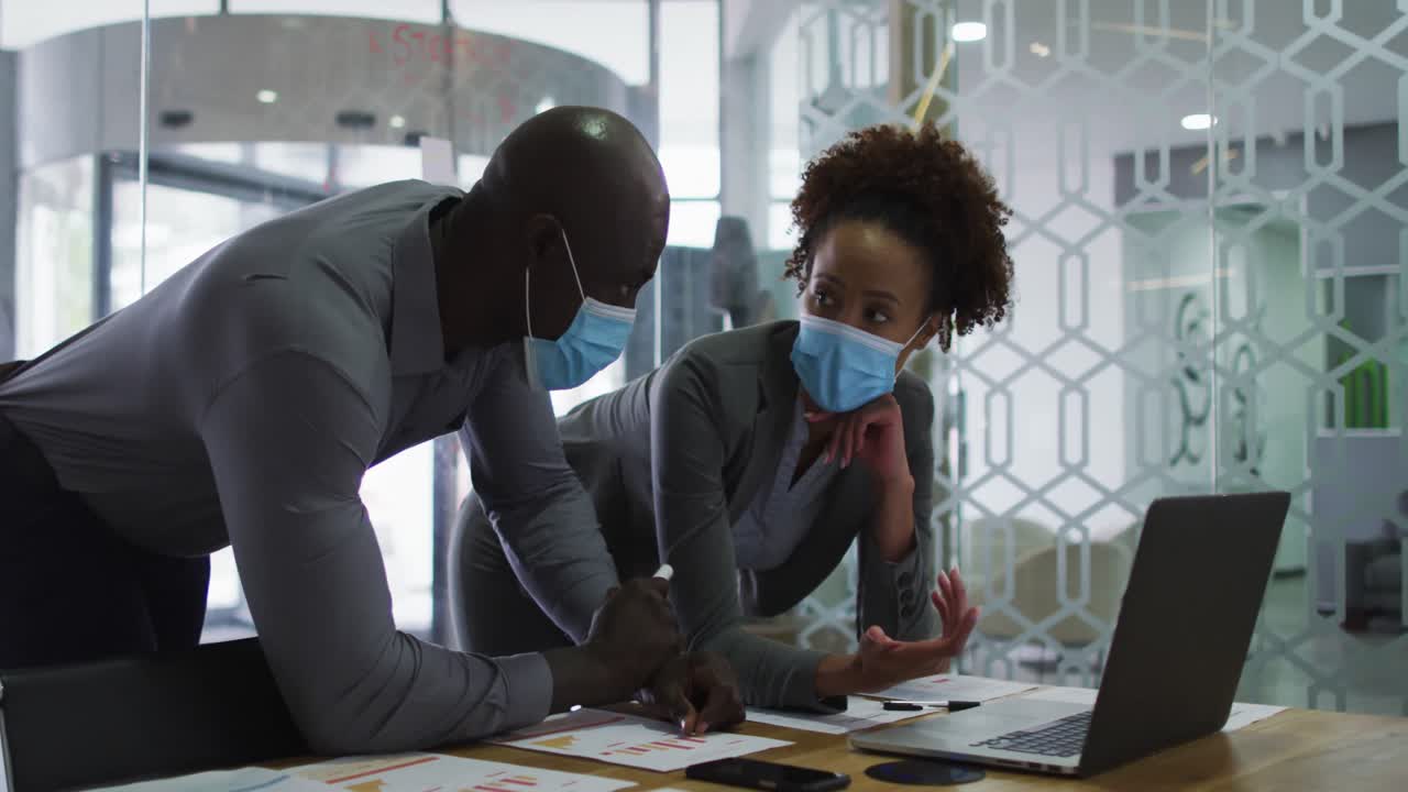 Diverse male and female business colleagues in face masks discussing, using laptop in office