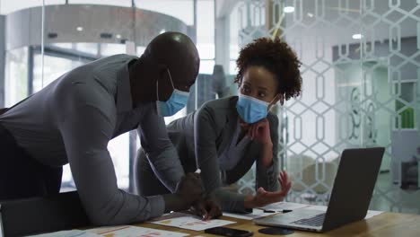diverse male and female business colleagues in face masks discussing, using laptop in office