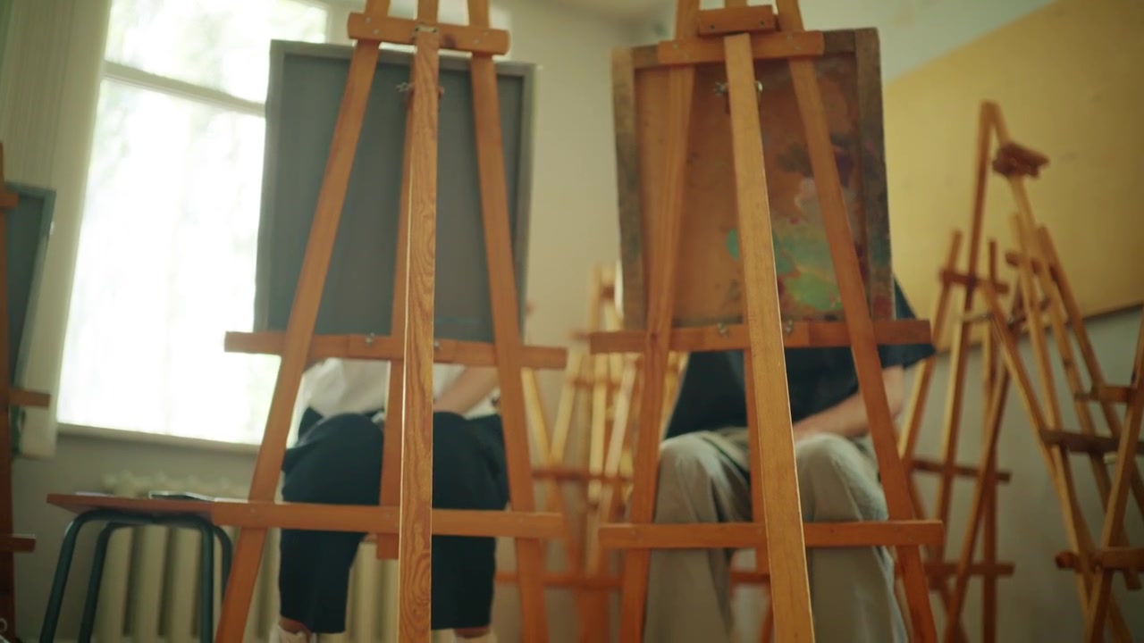 Cavalletti di legno disposti all'interno di una luminosa aula d'arte, che rappresentano l'educazione creativa, i laboratori artistici e gli spazi di apprendimento dedicati alle arti visive e al design.