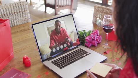 Happy-african-american-man-opening-gift-and-making-valentine's-day-video-call-on-laptop