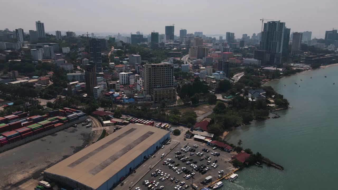 흐린 날 배경에 캄보디아 시아누크빌의 스카이라인이 있는 대형 창고 옆 대형 주차장에 여러 대의 자동차가 주차되어 있습니다.