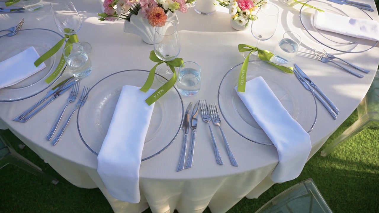 Beautifully arranged outdoor table features clear plates, white napkins, green ribbon accents, sparkling glassware, and pastel floral centerpieces under natural daylight. Perfect for weddings.