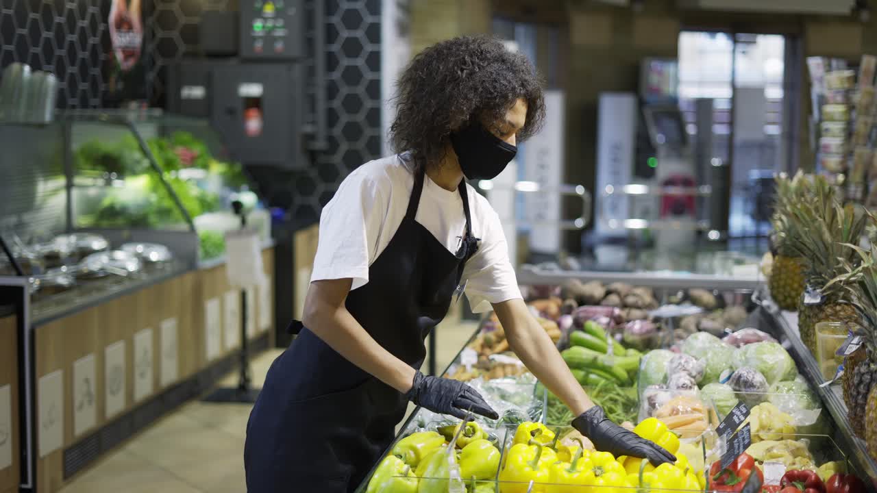 Funcionário de supermercado multirracial com máscara facial e luvas organizando vegetais e frutas frescas.