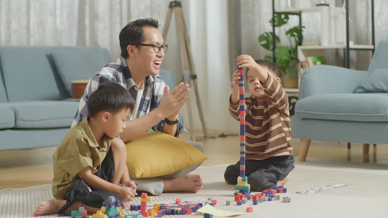 Full Body Of Asian Father And Sons Assemble The Construction Set Colorful Plastic Toy Brick On A Mat At Home. The Father Clapping Hands Celebrating Success, Another Arms Crossed Son Shaking Head Jealous His Brother