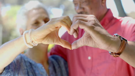 portrait of happy senior biracial couple making heart gesture in sunny garden at home, slow motion