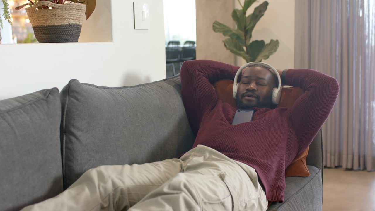 Uomo afroamericano che scorre lo smartphone, sistema le cuffie e si rilassa su un divano grigio a casa.