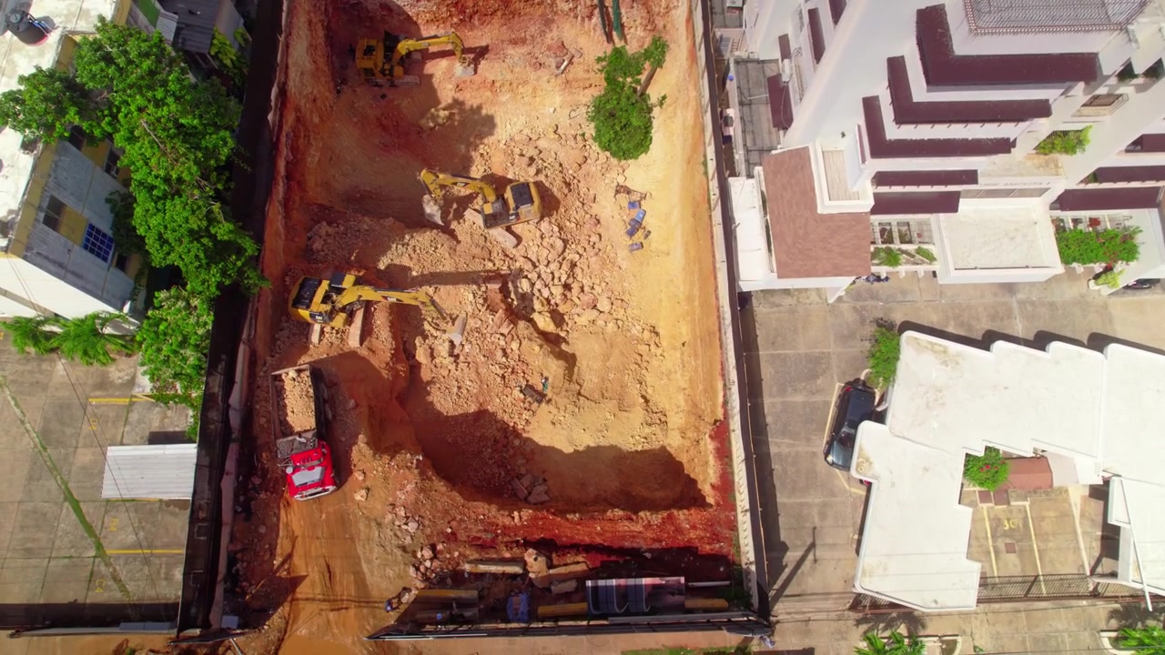 Tomada aérea de drone em ângulo elevado voando para frente e inclinando para baixo sobre árvores de parque para revelar um canteiro de escavação com máquinas pesadas para a construção de um novo edifício