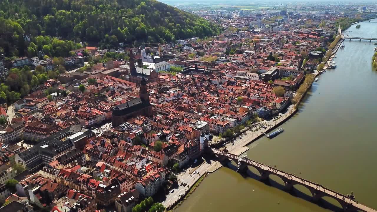 Aerial view of Heidelberg, Germany, showcasing its historic city center with red rooftops, iconic Old Bridge, Neckar River, and lush green hills surrounding the charming, well-preserved architecture