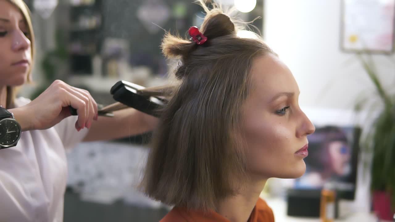 Side view footage of a beautiful young woman with a casual make up in beauty salon. Professional hairdresser styling her hair