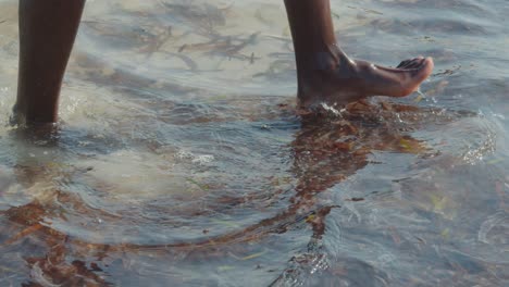 Las-Piernas-Y-Los-Pies-Desnudos-De-Un-Hombre-Negro-Caminando-En-El-Agua-Del-Océano-Con-Marea-Baja