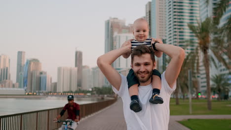 Lächelnder-Junger-Vater-In-Weißem-T-Shirt-Und-Hipster-Borsten,-Der-Mit-Seinem-Sohn-Auf-Dem-Hals-Am-Wasser-Sitzt,-Im-Hintergrund-Die-Moderne-Stadt-Im-Sommer-Am-Wasser