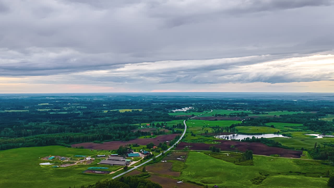 A wide aerial timelapse captures clouds moving over the vast Latvian countryside, showing a patchwork landscape of forests, farms, fields, lakes, and a winding country road