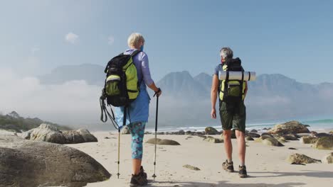 Vista-Trasera-De-Una-Pareja-De-Excursionistas-Mayores-Usando-Mascarilla-Con-Mochilas-Y-Bastones-De-Senderismo