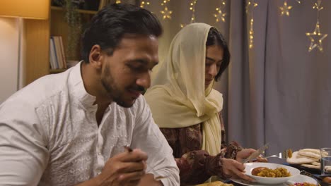 muslim family sitting around table at home with woman serving biryani at meal to celebrate eid