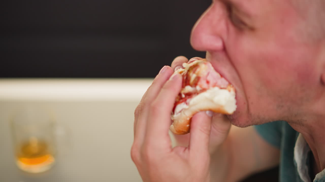 Close side view of man taking big bite of sausage sandwich indoors, holding sandwich with both hands, mouth wide open, juicy filling visible, with blurred background