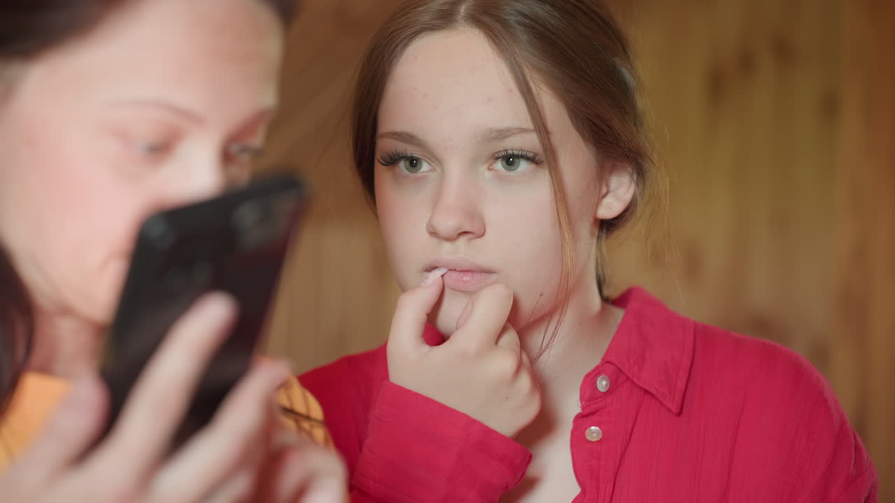 young girl in red shirt gazes pensively while watching friend in orange shirt operate smartphone in cozy wooden room, both engaged and focused on mobile content with thoughtful expressions