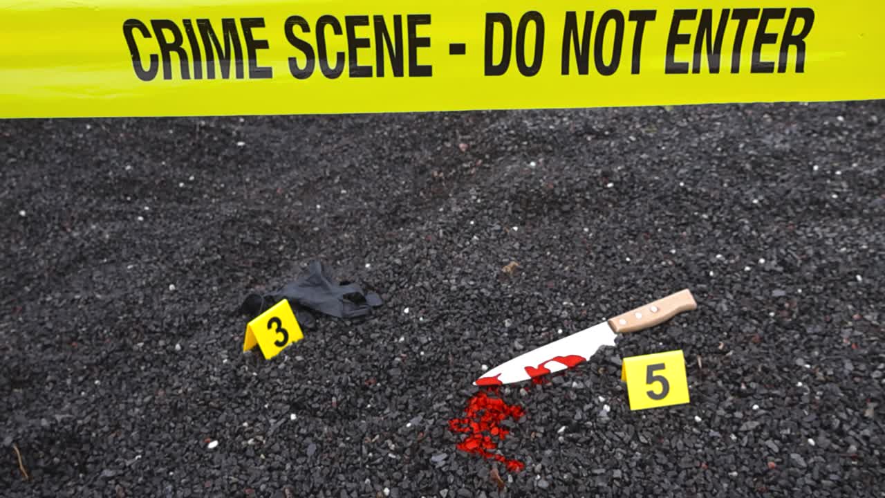 Police line or forensic investigation crime scene tape ribbon in front of a are where a black nylon glove and a bloody blood stained kitchen knife are on dark gravel outdoor at day time, bokeh