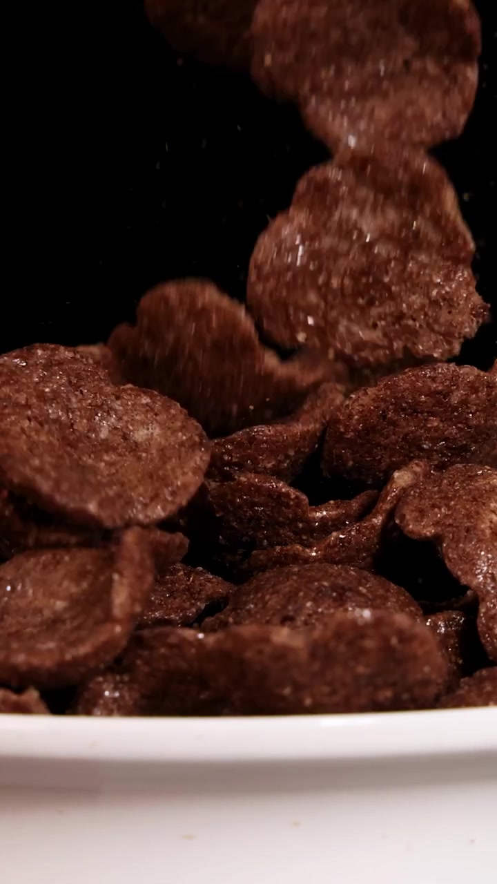Chocolate cereal pieces drop into a white bowl against a black background, creating a dynamic and appetizing scene