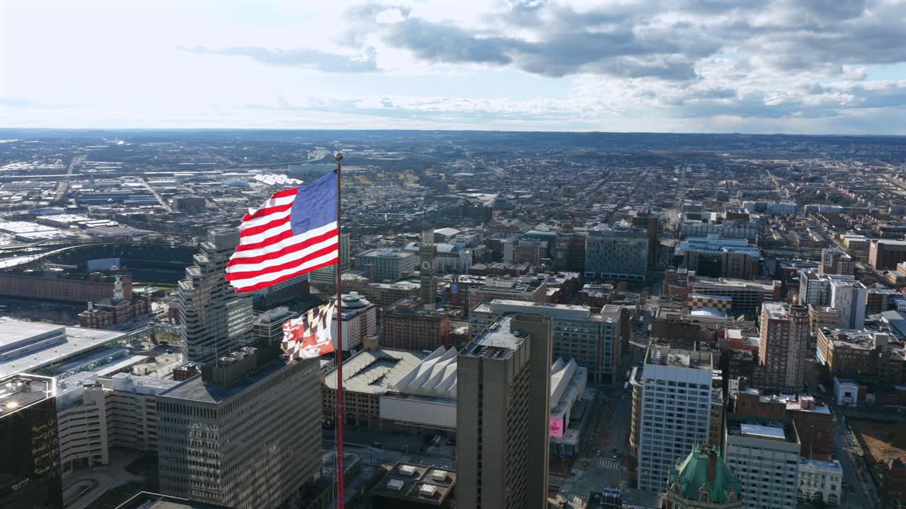 Vista aerea dello skyline di Baltimora con una grande bandiera americana che si erge sopra gli edifici del centro, che mostra una fitta architettura urbana sotto nuvole suggestive in pieno giorno. Bandiera degli Stati Uniti e del Maryland in orbita