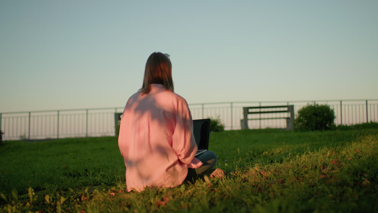 핑크색 을 입은 프리랜서의 뒷면, 안경이 머리에 앉아 있으며, 노트북에 집중하고, 태양 그림자가 옷에 패턴을 만들고, 먼 배경의 벤치