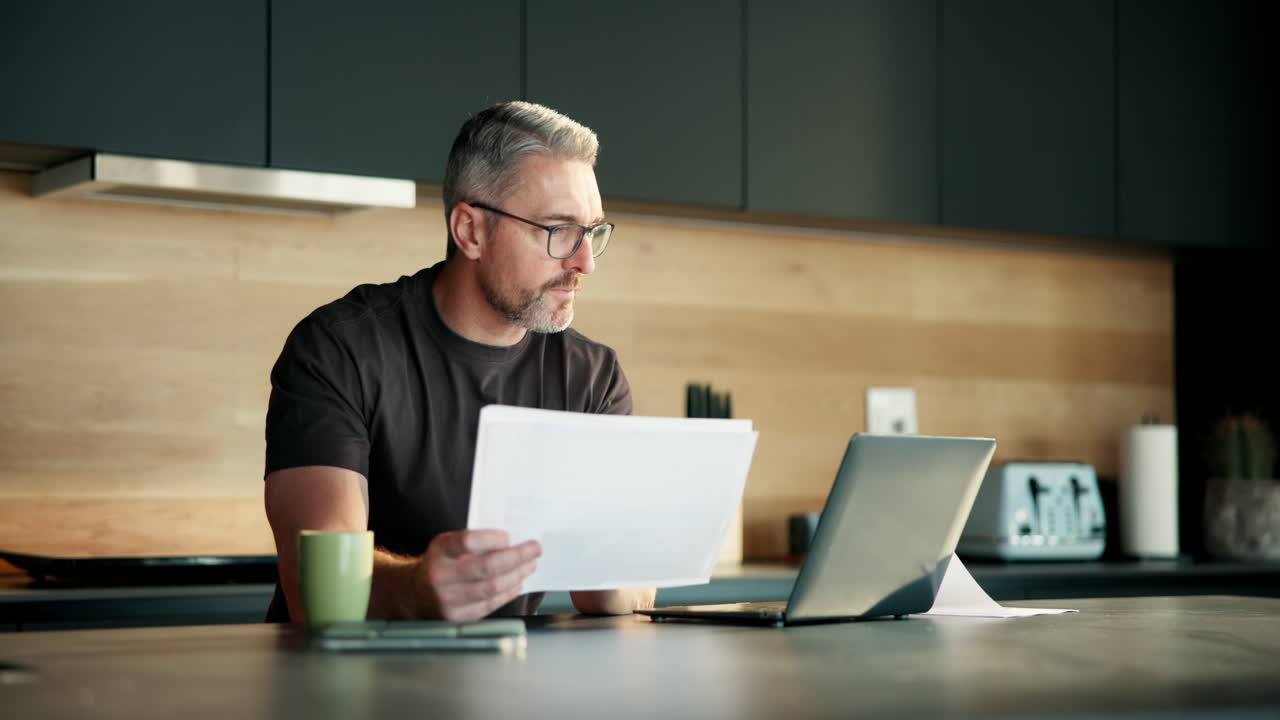 Uomo concentrato che legge documenti e lavora al laptop in una cucina moderna