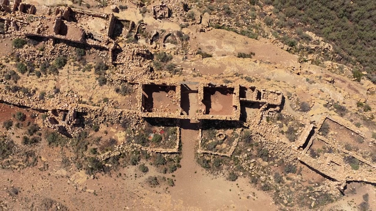 Uma vista aérea de drone de cima para baixo revela os padrões geométricos de ruínas de pedra abandonadas no Sudoeste. As paredes desgastadas e as estruturas sem telhado contrastam fortemente com o chão árido do deserto.