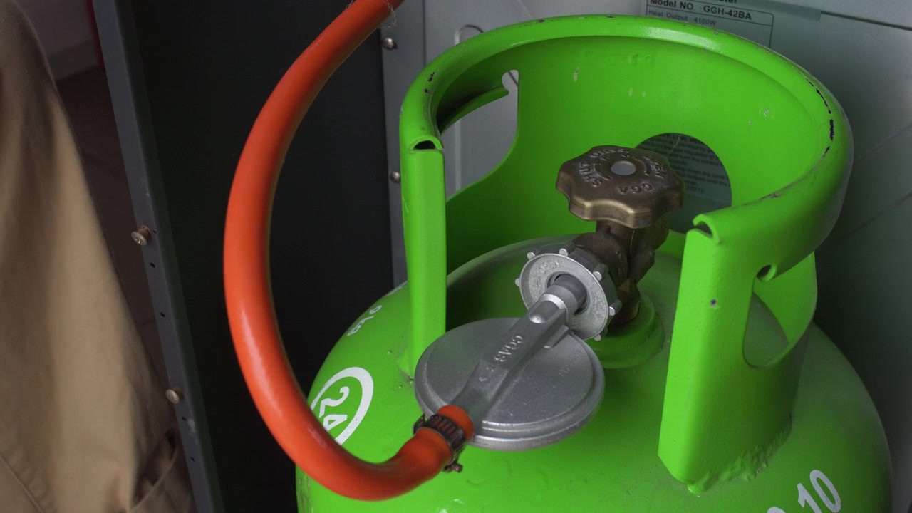 Hænder placerer en sølvfarvet reguleringsventil og skruer den stramt fast på messingdysen på en grøn LPG-gasflaske. Statisk nærbillede
